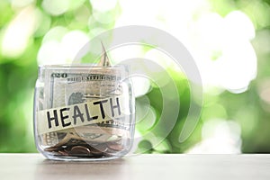 Glass jar with money and label HEALTH on table against blurred background.