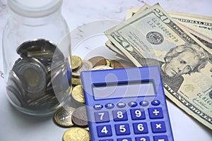 Glass jar with multicurrency coins, US dollar bills and calculator white table