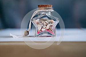 A glass jar in the shape of a star inside pink sand and small shells on a blue background close-up.Maritime concept