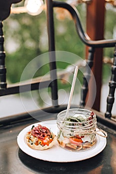 Glass jar with snacks and tartlet with tomatoes and meat on a white plate close-up