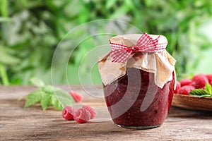 Glass jar of sweet jam with ripe raspberries on wooden table against blurred background.