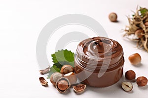 Glass jar with tasty chocolate spread, hazelnuts and green leaves on light background