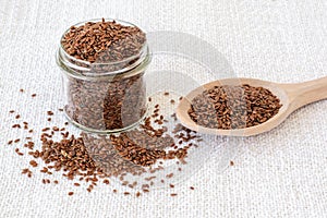 Glass jar and wooden spoon are full of whole flax seeds on a white textile table mat. Healthy food
