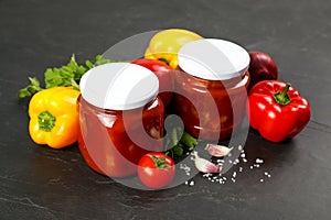 Glass jars of delicious canned lecho and fresh ingredients on black table