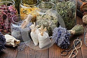 Glass jars of dry different medicinal herbs, aromatic sachets, bunches of dry lavender on table.
