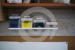 Glass jars filled with pigments in blue, yellow, and teal rest on a workshop shelf, hinting at the vibrant creations