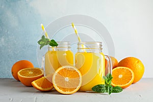 Glass jars with fresh orange juice and tubule, oranges and mint on white table against color background, space for text