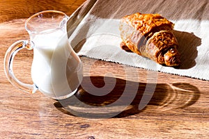 Glass jug of milk with croissant on wooden table