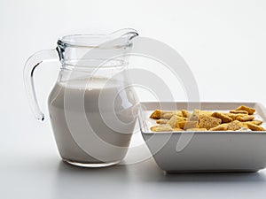A glass jug with milk next to a square bowl with cereals
