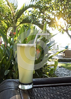 A glass of juice on the edge of the table on the street terrace, overlooking the pool and tropical plants. Indonesia, Bali