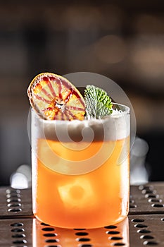 Glass of Jungle Bird cocktail garnished with dried red orange and mint leave on bar counter