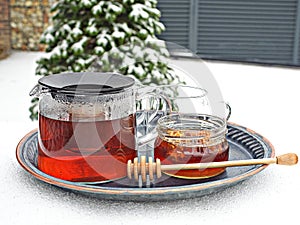 Glass kettle of hot tea and jar of honey on table covered by snow