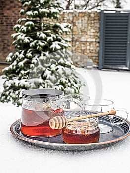 Glass kettle of hot tea and jar of honey on table covered by snow