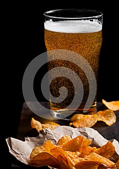 Glass of lager beer with potato crisps snack on vintage wooden board on black background. Beer and snack