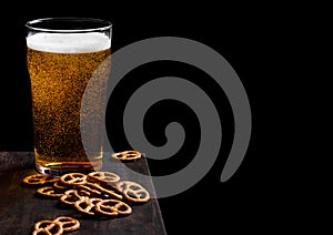 Glass of lager beer with pretzel snack on vintage wooden board on black background. Beer and snack. Space for text