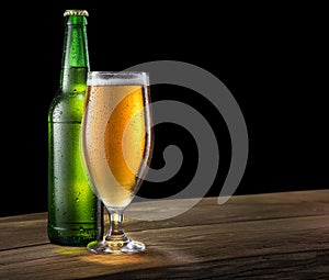 Glass of light beer and green beer bottle on the bar counter on a black