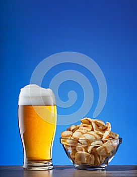 Glass of light beer and plate of chips on blue background