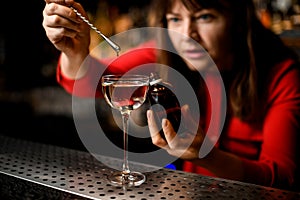 glass with liquid stands on bar and woman adds ingredient to it