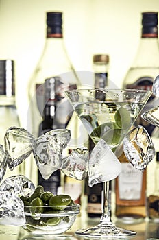 Pieces of ice, glass bowl with green olives and a glass of martini