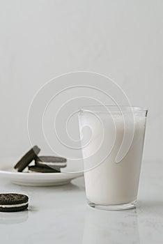 Glass of milk accompanied by some chocolate cookies filled with white cream on a white background