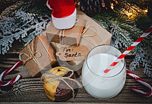 A glass of milk, cookies, santa hat and gifts on the table.
