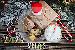 A glass of milk, cookies, santa hat, numbers 2022, an inscription from letters: xmas and gifts on the table.