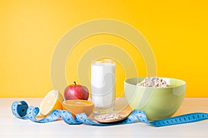 glass of milk, fruits, a bowl of oatmeal and a blue measuring tape on the table, yellow background, healthy lifestyle concept