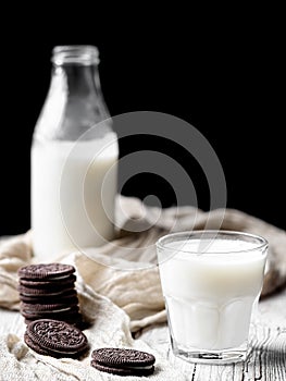 A glass with milk and several chocolate cookies on a wooden surface. Close-up. Bottle of milk in the background. Natural dairy