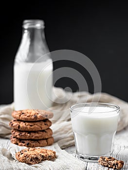 A glass with milk and several oatmeal cookies with chocolate on a wooden surface. Close-up. Bottle of milk in the background.