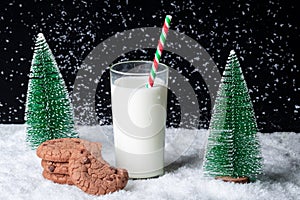 A glass of milk with a straw and a half-eaten cookie on a snowy night background among toy Christmas trees
