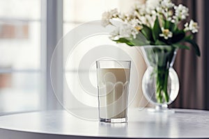 glass of milk on table and vase with flowers and window as background