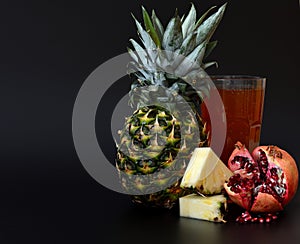 A glass of a mixture of freshly squeezed exotic fruits on a black background, next to a pineapple fruit and a broken ripe