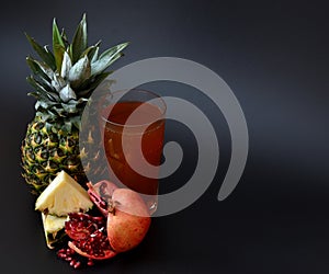 A glass with a mixture of tropical fruit juices on a black background, next to pieces of ripe pineapple and a broken pomegranate