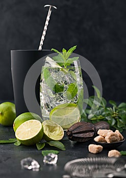 Glass of Mojito cocktail with ice cubes mint and lime on black board with spoon in shake and wooden squeezer and fresh limes