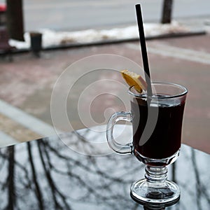 Glass mug with brown, tasty, hot, fragrant, black tea with a slice of lemon and a straw on the table in a cafe in the evening in t
