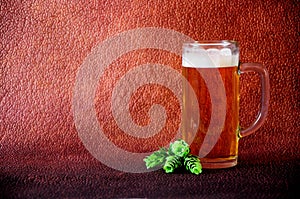 Glass mug with a handle full of light beer with white foam and hop fruit on a brown abstract background
