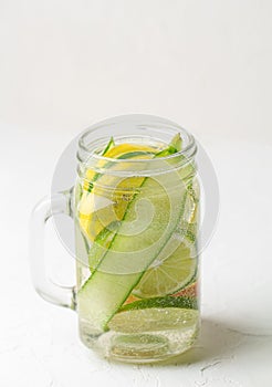 Glass mug with handle with refreshing detox lemonade with sliced lime, lemon, cucumbers and red orange on white table.