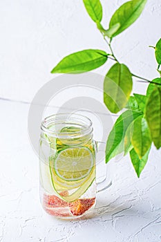 Glass mug with handle with refreshing detox lemonade with sliced lime, lemon, cucumbers and red orange on white table.