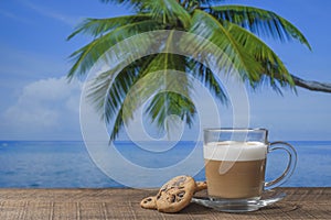 Glass mug of hot cappuccino coffee with cookies on a wooden table with sea water, coconut palm tree and blue sky background on