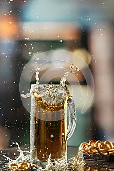 Glass mug of light cold beer with splash and flying drops with snacks on the table in pub