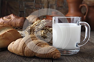 Glass mug with milk, bun and bakery products lying on an old table on the background of wheat spikes and ceramic dishes. Close-up.