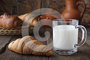 A glass mug with milk and a fresh bun lying on an old table on a background of baking from wheat flour and ceramic dishes. Close-u