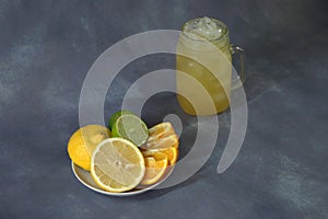 A glass mug with a refreshing juice with ice and a plate with citrus platter, lemon, lime and orange, whole and cut into pieces.