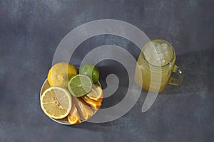 A glass mug with a refreshing juice with ice and a plate with citrus platter, lemon, lime and orange, whole and cut into pieces.