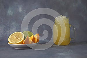 A glass mug with a refreshing juice with ice and a plate with citrus platter, lemon, lime and orange, whole and cut into pieces.