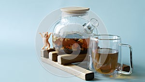 Glass mug and teapot with hot black flavored tea with fruits and flower petals on a wooden hot plate. Next to a toy dancing kitten