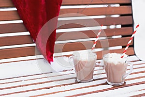 Glass mugs, cups with hot chocolate, cocoa, marshmallows. Santa Claus hat. Winter snow covered park bench. Striped red paper