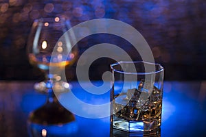 Glass o whisky with ice and wineglass of cognac on yellow-blue bokeh background