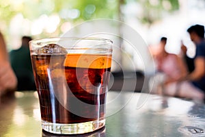 Glass ofdark spanish vermouth drink with slice of orange, a popular aperitif, on outdoor bar table with people during summer day