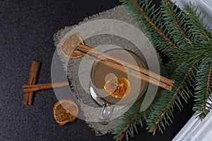 A glass of orange drink stands on a piece of linen. A slice of dried orange floats in it. Nearby are cinnamon sticks and dried